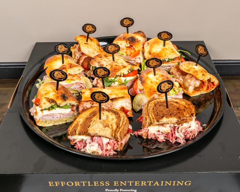 A black platter filled with various sandwiches, including Reubens, meatball subs, and Italian subs.