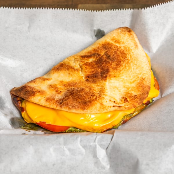 A grilled flatbread sandwich with melted yellow cheese, red tomato, and green pesto on white parchment paper.