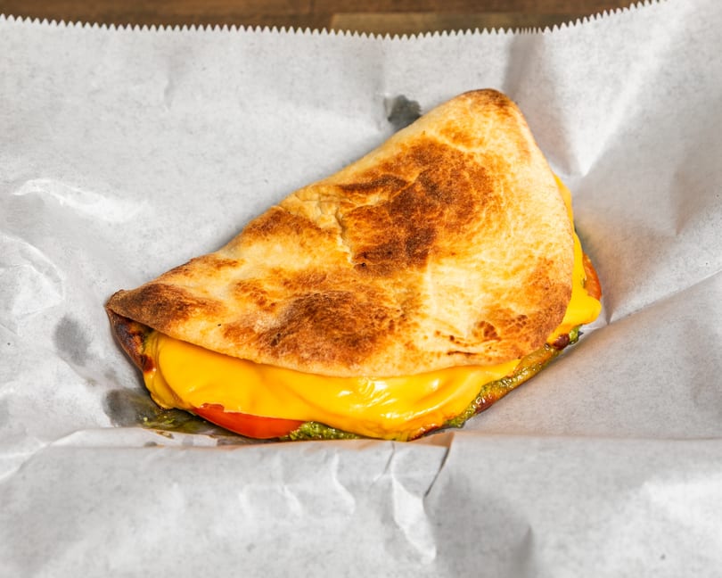 A grilled flatbread sandwich with melted yellow cheese, red tomato, and green pesto on white parchment paper.