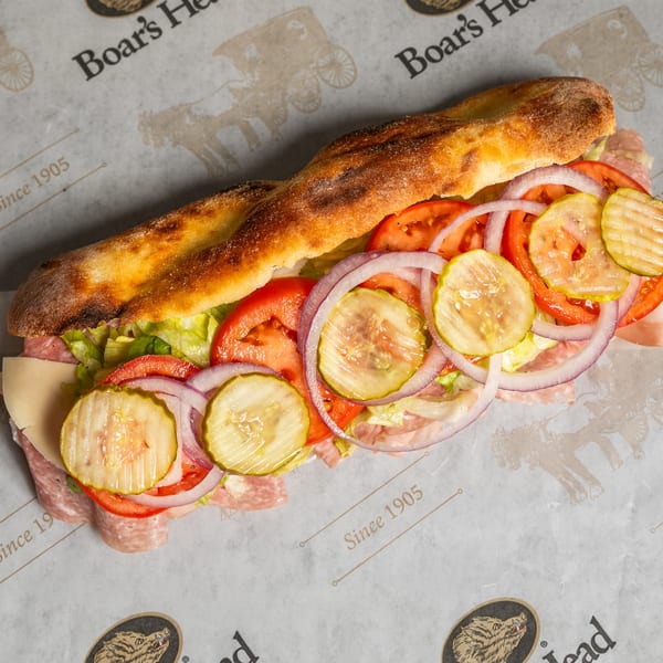 A Boar's Head sub sandwich with salami, cheese, lettuce, tomatoes, onions, and pickles on branded paper.