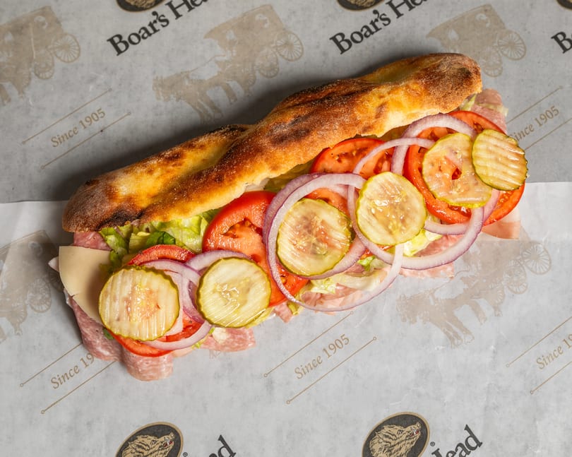 A Boar's Head sub sandwich with salami, cheese, lettuce, tomatoes, onions, and pickles on branded paper.