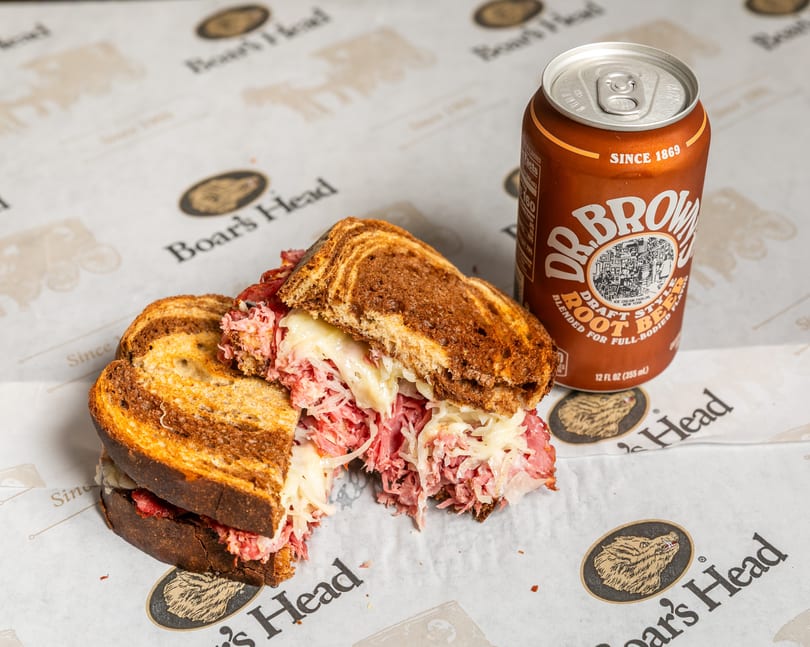 A Boar's Head Reuben sandwich with melted cheese and sauerkraut, next to a Dr. Brown's Root Beer.