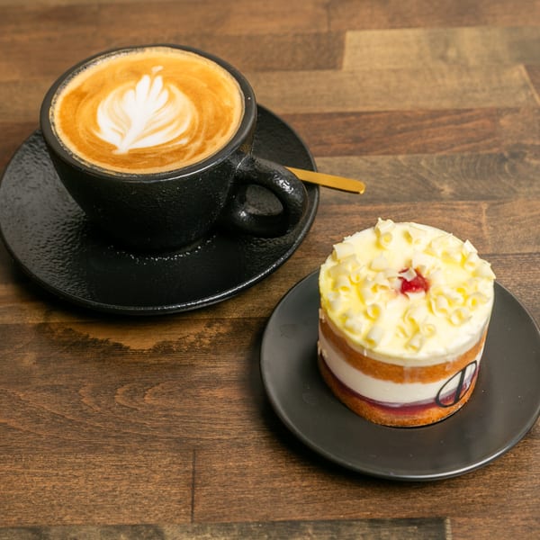Latte with latte art and a layered cake with white frosting and chocolate curls on a wooden table.