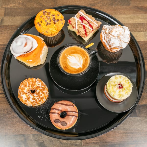 A black tray with a latte, cinnamon roll, two donuts, a muffin, a crumble bar, and a small cake.