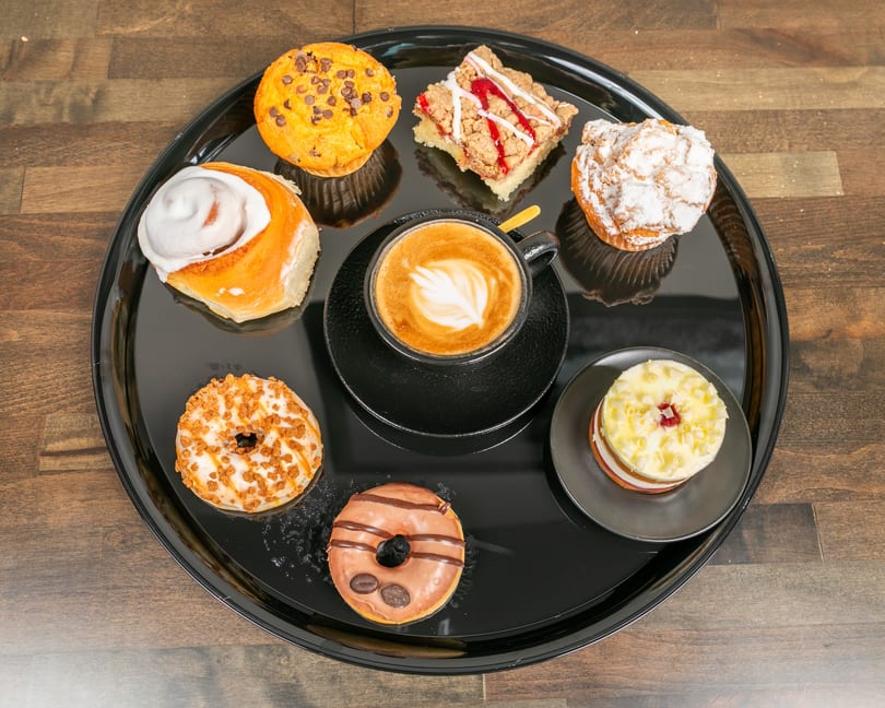 A black tray with a latte, cinnamon roll, two donuts, a muffin, a crumble bar, and a small cake.